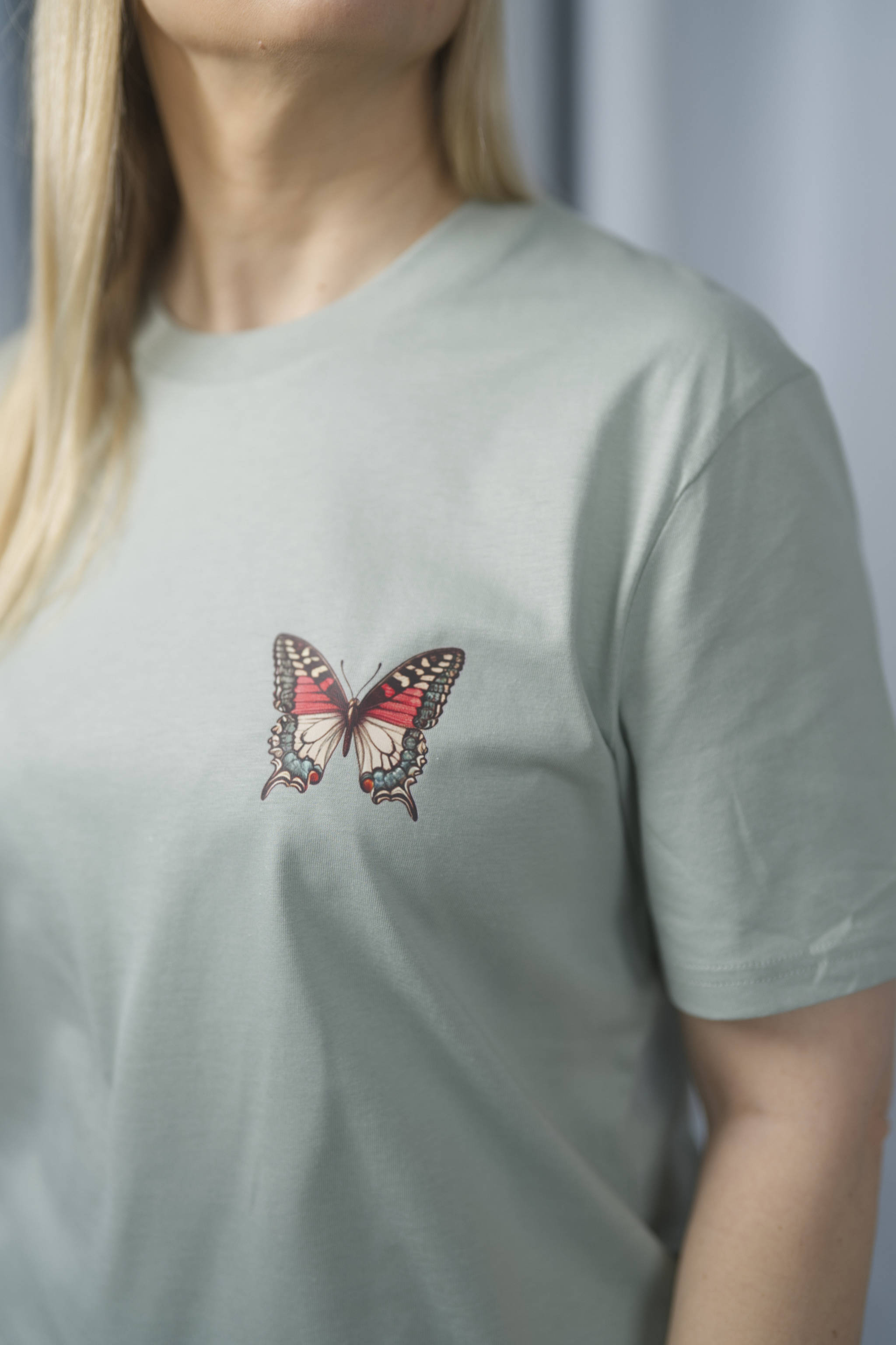 T-Shirt aus 100 % Bio-Baumwolle mit normaler Passform, erhältlich in verschiedenen Farben wie Blue Soul, Cotton Pink, Lavender, Aloe, Weiss und Schwarz. Individuell bedruckbar mit Wunschmotiv. Weiches, angenehm leichtes Material für hohen Tragekomfort. Ideal für einen zeitgemässen, entspannten Look. Vorderseite: Farbiger Schmetterling auf der linken Brust. Rückseite: Buntes Blumen- und Schmetterlingsfeld im Illustrationsstil.