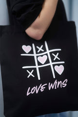 Schwarze Tote Bag mit einem verspielten Tic-Tac-Toe-Design aus weissen Linien und rosa Herzen sowie dem pinken Schriftzug „LOVE WINS“ – stilvolle Stofftasche mit Statement für Liebe und Vielfalt.