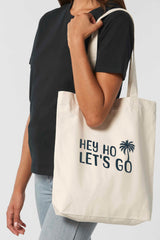 Naturfarbene Tote Bag mit schwarzem Schriftzug „HEY HO LET’S GO“ und Palmenmotiv – nachhaltige Stofftasche aus recycelten Materialien mit langen Henkeln und geräumigem Schnitt.